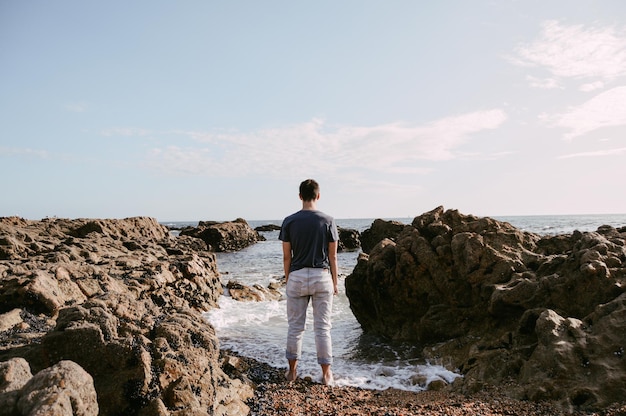 rear view of young man standing at rocky shore against sky