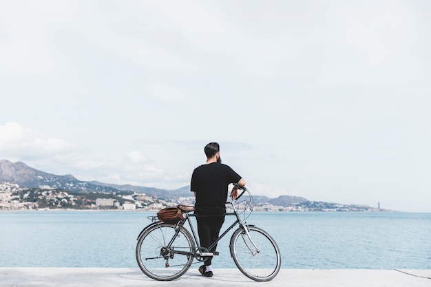 Rear view of a young man standing with bicycle looking at sea