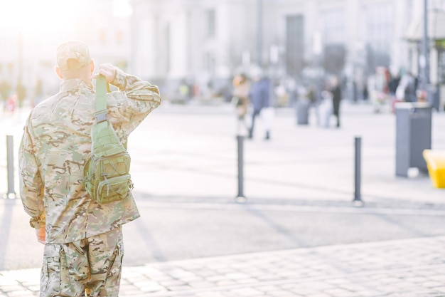 Rear view of a young military man in a camouflage uniform and a cap standing on the street near the station waiting for relatives and friends The soldier returned from the army