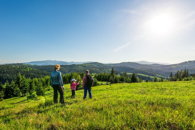 背面図若い母親と2人の娘は、緑の草が生い茂った丘を下ります。暖かい夏の日のトレッキング中に山に生えている森のゴージャスな景色