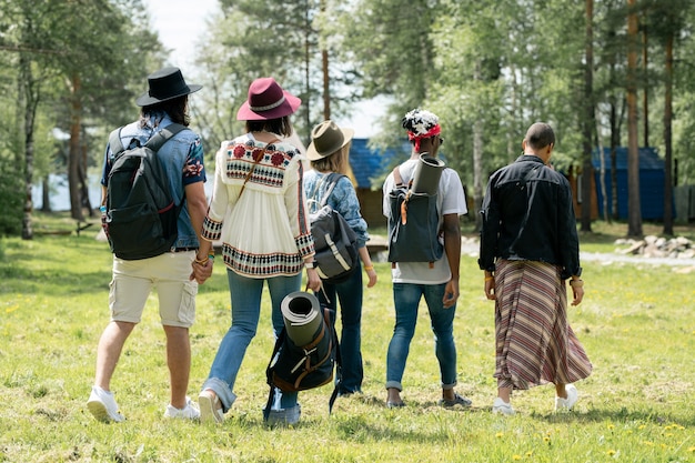 Vista posteriore di giovani amici multietnici con cartelle che camminano sul campeggio del festival mentre trovano posto per il campeggio