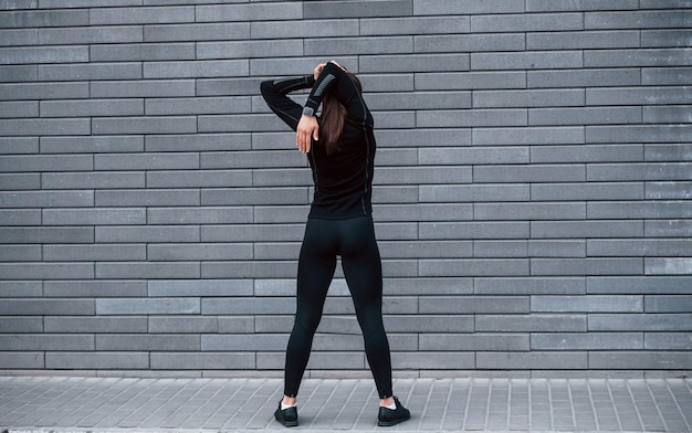 야외 회색 벽 근처에 서 있는 검은색 운동복을 입은 낚시를 좋아하는 어린 소녀의 뒷모습
