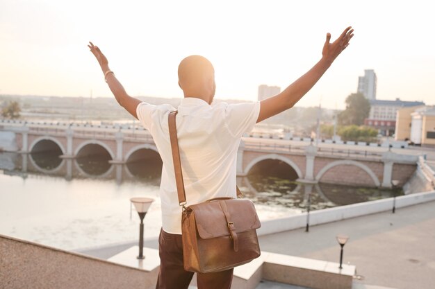 Vista posteriore del giovane uomo d'affari africano vincente con borsa in pelle che esprime trionfo di fronte al fiume e all'architettura moderna