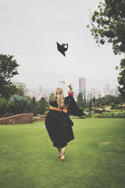 Photo rear view of young woman in graduation gown throwing mortarboard while walking on grassy field at park