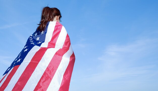 Rear view of a young woman holding an American flag on her shoulders against a blue sk