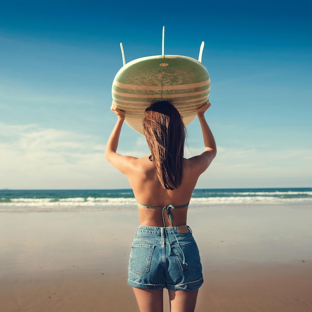 Photo rear view of young woman holding surfboard while standing at beach against sky