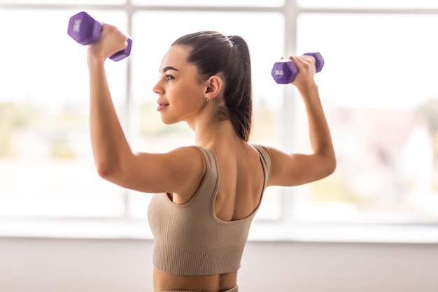 Photo rear view of young woman in modern light beige closefitting sportswear holding blue dumbbells and showing fit body