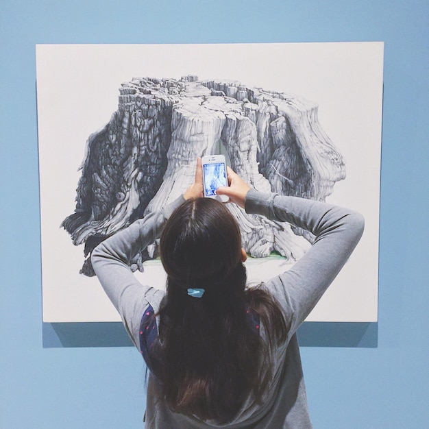 Foto vista posteriore di una giovane donna che fotografa un dipinto attraverso uno smartphone