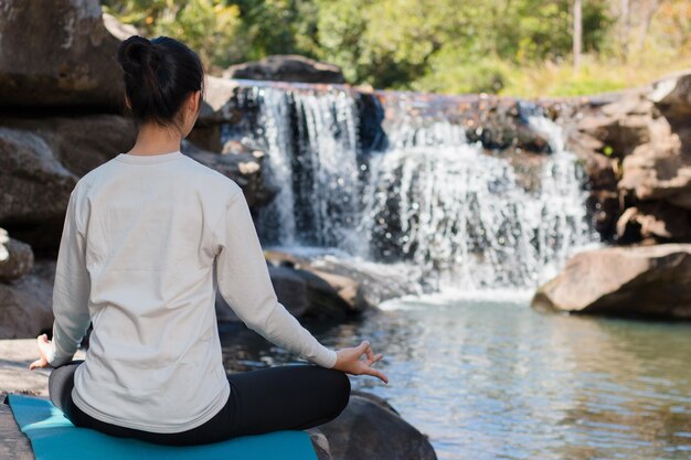 Rear view of young woman practicing yoga on rock against waterfall in forest