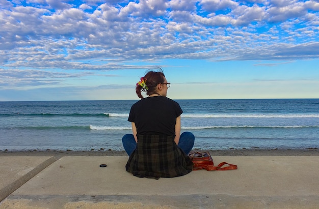 Rear view of young woman sitting on shore at beach against sky during sunset