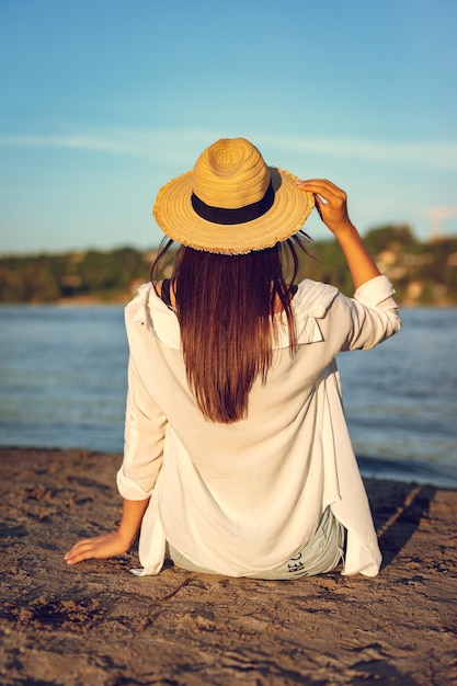 川岸で日没時にリラックスした麦わら帽子の若い女性の背面図。
