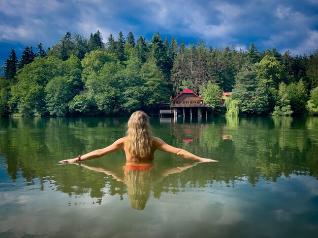 Foto vista posteriore di una giovane donna che nuota in un lago circondato da una foresta verde in una nuvolosa giornata estiva