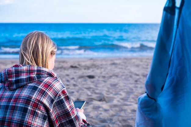 Photo rear view of young woman using mobile phone while sitting at beach against sky