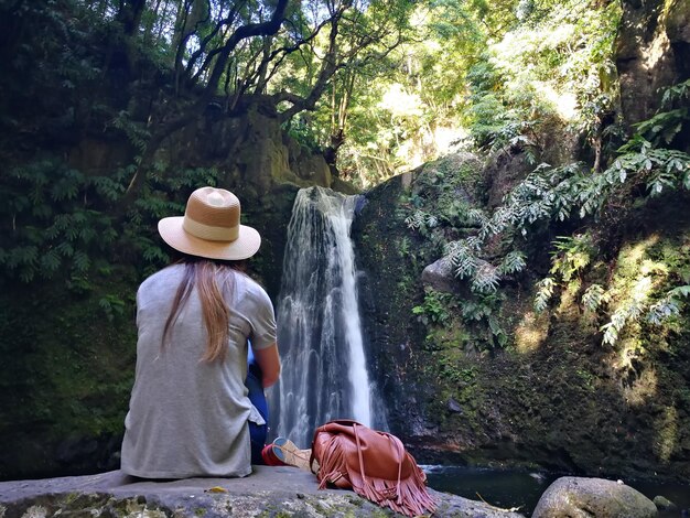 Photo rear view of young woman wearing hat looking at waterfall while sitting on rock in forest