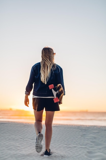 夕暮れの晴れた空に沿ってスケートボードでビーチを歩く若い女性の後ろの景色