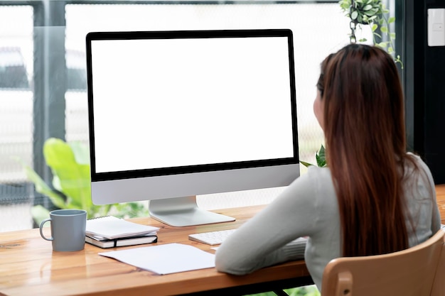 Rear view of young woman working on desktop computer at home, mockup blank screen monitor of product display or graphic design.