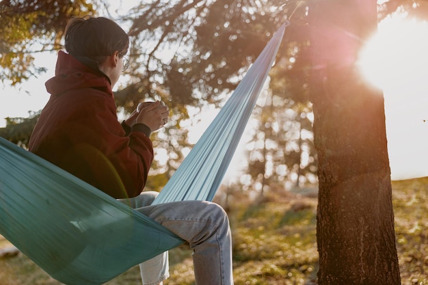 Photo rear on woman with short hair holding thermos of coffee and resting in hammock in wood