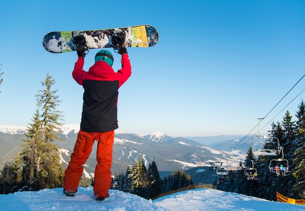 Colpo integrale retrovisore di uno snowboarder in piedi in montagna alzando il suo snowboard in aria sopra la sua testa copyspace felicità godimento ricreazione sport invernali concetto