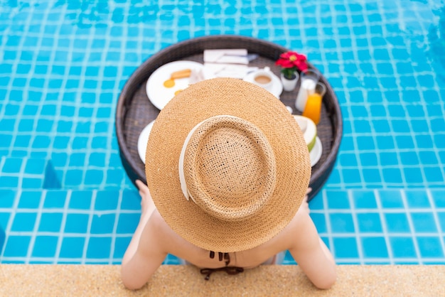 Rearview of a woman in sun hat relaxing in a pool with a floating breakfast tray at a luxury hotel