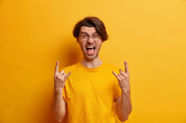 Rebelious unshaven hipster guy makes rock gesture screams out loudly being part of concert band wears round glasses and casual t shirt isolated over yellow  wall. Body language concept