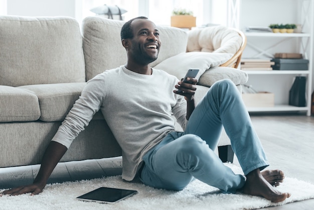 Receiving funny messages. Young African man holding a phone and smiling while sitting on the carpet at home