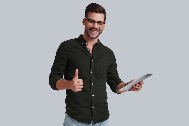 Receiving good feedbacks. Good looking young man in eyeglasses holding his digital tablet and gesturing while standing against grey background