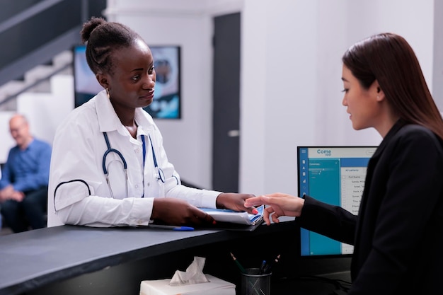 Photo receptionist helping physician doctor with checkup visit appointments while working at hospital registration desk in waiting area. medic reviews the patients medical history before their consultation.