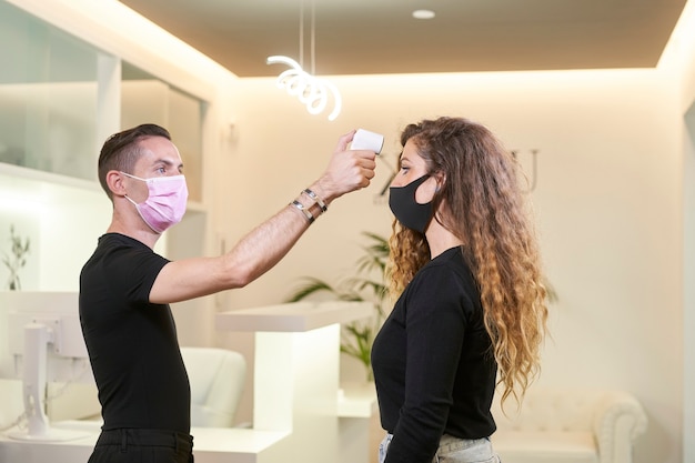 Receptionist using an infrared thermometer to take temperature. Female patient wearing medical mask in examination room at clinic.