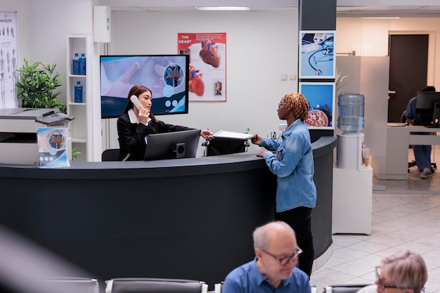 Receptionist using landline phone at reception desk, giving\
report papers to patient to fill in before medical checkup\
appointment. worker helping woman with consultation forms, cord\
telephone.