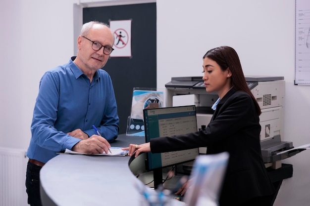Receptioniste die verzekeringspapier laat zien aan oude patiënt en hem laat ondertekenen voordat hij begint met consultatie tijdens controlebezoek in de ziekenhuislobby. Geneesmiddelen worden gebruikt om symptomen te verlichten en de gezondheid te verbeteren