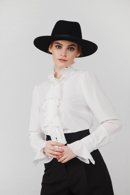 Reclame stijlvolle schattige jonge zakenvrouw in zwarte elegante blouse en hoed op witte lichte achtergrond. Vrouw in modieuze kleding poseren. Concept van stijl, mode, schoonheid en het bereiken van doelen