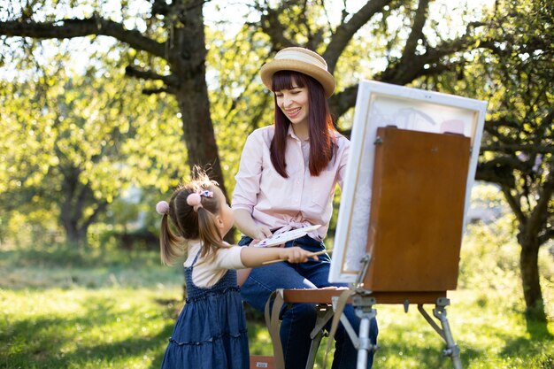 Recreation and family summer games in the open air. Young laughing mother enjoying her joint time with little daughter, teaching painting with easel in garden.