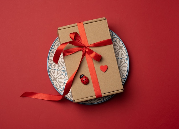 Прямоугольная коричневая картонная коробка, перевязанная красной шелковой лентой, лежит на круглой керамической красной тарелке, вид сверху