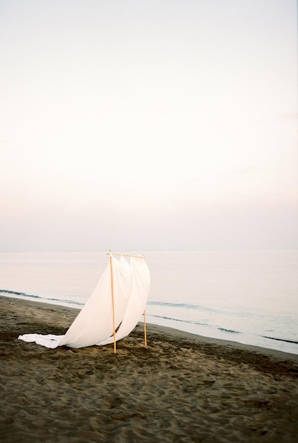 Rectangular wedding arch with white fabrics fluttering in the wind stands on a sandy beach