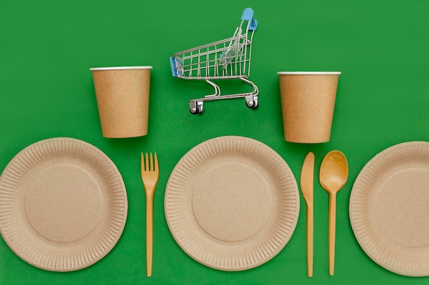 On a recyclable plate is a small shopping cart, next to a fork, spoon, knife, and a cup on a green background. Kitchen utensils are served on the table. Top view. Minimalist Style. Copy space