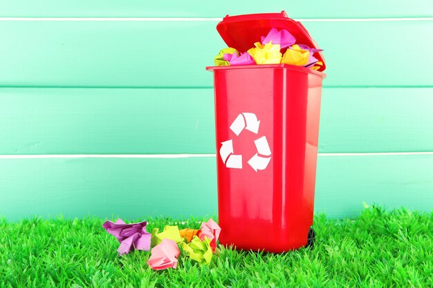 Recycling bin with papers on grass on light blue background