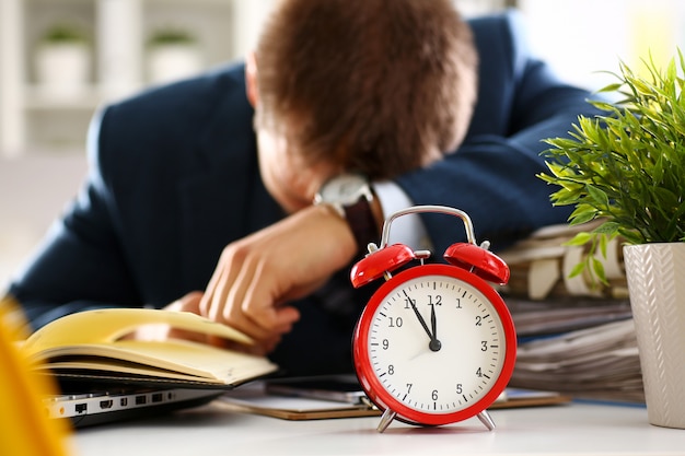 Red alarm clock shows late time closeup and tired office male clerk in suit take nap on table workplace full of exam papers.