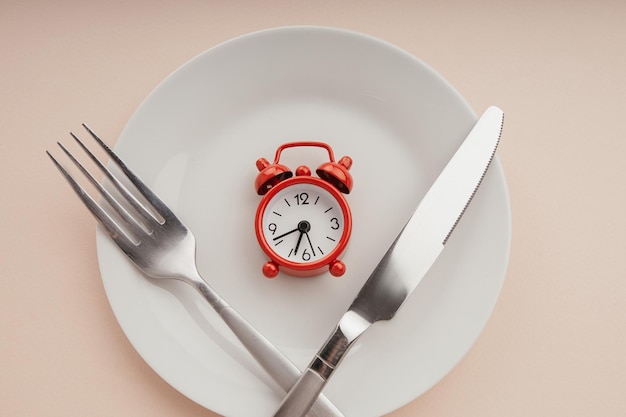 Red alarm clock on white plate with cutlery Concept of intermittent fasting lunchtime diet and weight loss