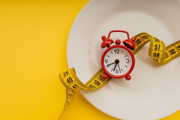 Red alarm clock on white plate with measuring tape on a yellow background closeup