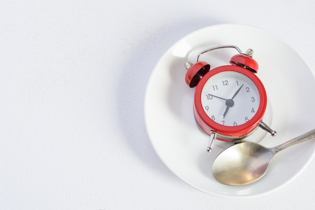 Red alarm clock on white plate with a silver spoon