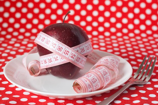 Red apple wrapped in a tape measure lying on a white plate on a red polka dot