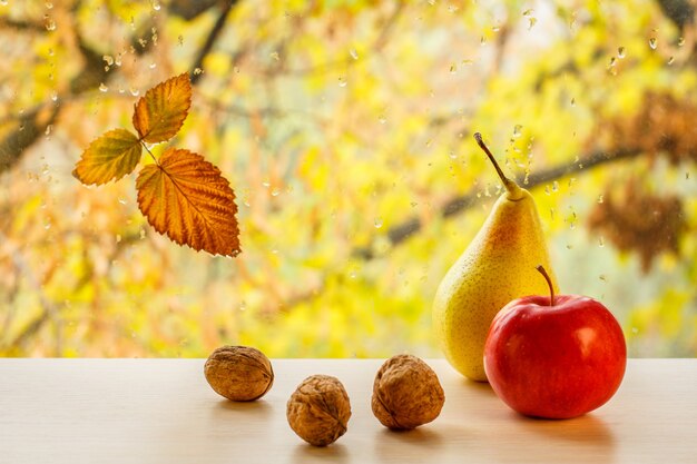 Красное яблоко, желтая груша, грецкие орехи и сухой лист на оконном стекле с каплями воды на размытом естественном фоне. Опавшие листья и капли дождя на оконном стекле с осенними деревьями на заднем плане.