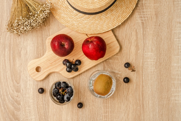 Red apples and a bunch of black grapes on wooden board , a bunch of black grapes and kiwi fruit in the glass, straw hat and grass flowers on wooden table surface