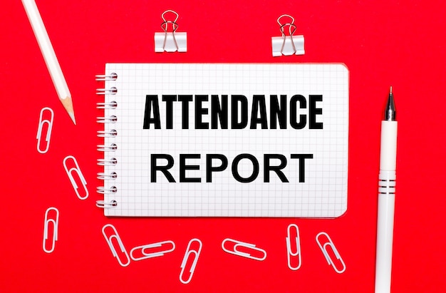 On a red background, a white pen, white paper clips, a white pencil and a notebook with the text ATTENDANCE REPORT. View from above