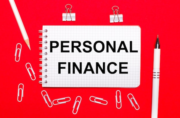 Photo on a red background, a white pen, white paper clips, a white pencil and a notebook with the text personal finance. view from above