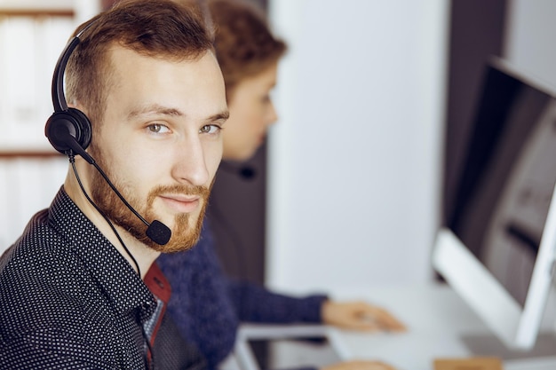 Red-bearded businessman talking by headset near his female colleague while sitting in modern sunny office. Diverse people group in call center.