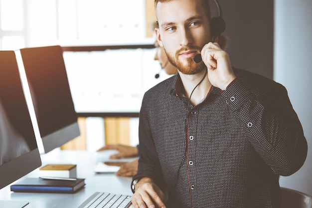 Red-bearded businessman talking by headset near his female colleague while sitting in sunny office. Diverse people group in call center.