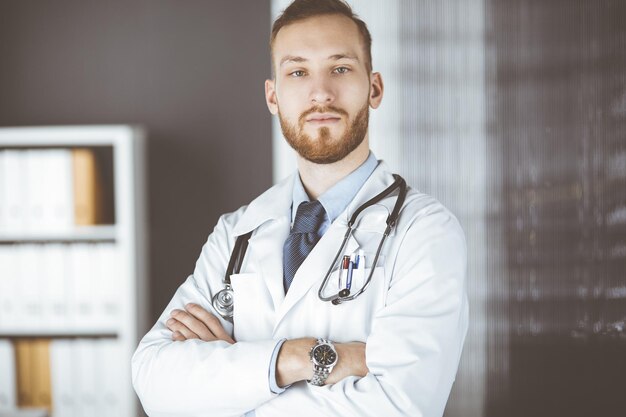 Red-bearded doctor standing straight in clinic near his working place. Portrait of physician. Medicine and healthcare concept.