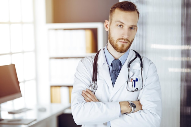 Red-bearded doctor standing straight in sunny clinic near his working place. Portrait of physician. Medicine concept.