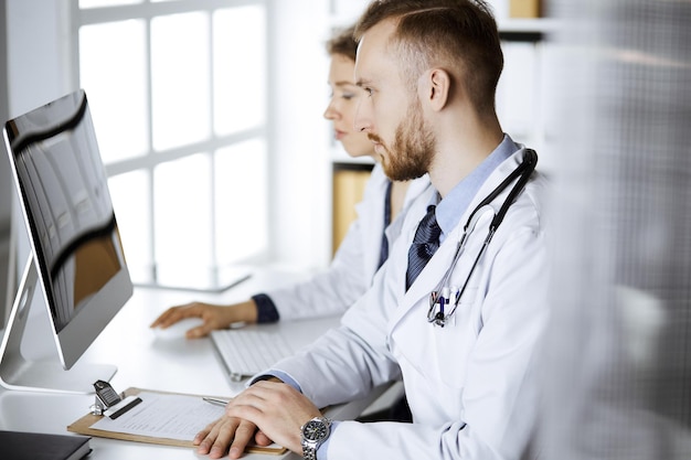 Red-bearded doctor with female colleague discussing current disease therapy while sitting at working place in clinic. Team work in medicine.